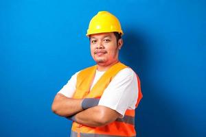 retrato de trabalhador asiático gordo usando capacete e colete de segurança olhando para a câmera cruzou a mão com gesto de confiança contra fundo azul foto