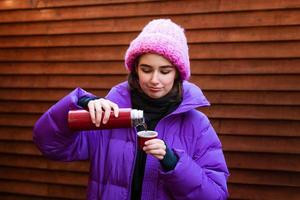 jovem feliz. uma mulher está bebendo chá quente enquanto segura uma garrafa térmica em um inverno foto