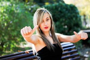 menina bonita sentada em um banco no parque mostrando o polegar para cima o sinal foto