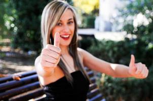 menina bonita sentada em um banco no parque mostrando o polegar para cima o sinal foto