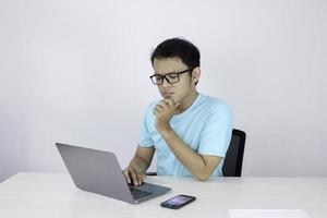 jovem asiático é sério e se concentra ao trabalhar em um laptop em cima da mesa. homem indonésio vestindo camisa azul. foto