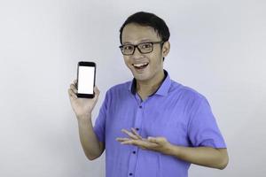 jovem asiático usa camisa azul está de pé e sorrindo apontando no espaço em branco na tela do smartphone em fundo branco. foto