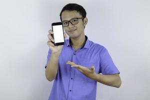 jovem asiático usa camisa azul está de pé e sorrindo apontando no espaço em branco na tela do smartphone em fundo branco. foto