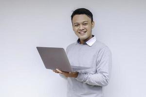 jovem asiático se sentindo feliz e sorrindo ao ficar de pé e trabalhar no laptop. homem indonésio vestindo camisa cinza foto