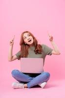 jovem mulher asiática sentada e usando laptop em fundo rosa foto