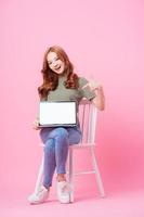 jovem mulher asiática sentada e usando laptop em fundo rosa foto
