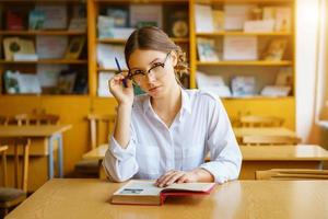 menina de óculos sentada em uma mesa com um livro na sala de aula, segurando um lápis na mão, olhar pensativo foto