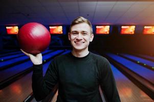 alegre jovem segurando uma bola de boliche e sorrindo para a câmera em pé contra pistas de boliche com luz ultravioleta. foto