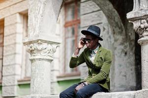 retrato da moda do homem afro-americano negro na jaqueta de veludo verde e chapéu preto, sentado na grade e falando em seu fundo de telefone celular antiga casa vintage com arco foto