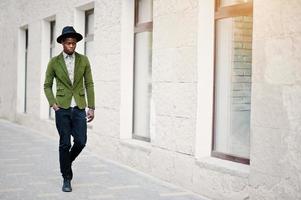 retrato da moda do homem afro-americano negro na jaqueta de veludo verde e chapéu preto andando nas ruas da casa de fundo da cidade com muitas janelas foto