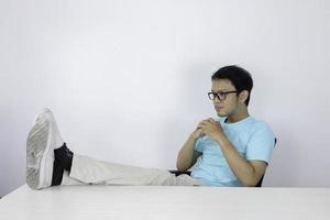 jovem asiático com olhar sério e de foco com a perna na mesa e o braço no queixo. homem indonésio vestindo camisa azul. foto