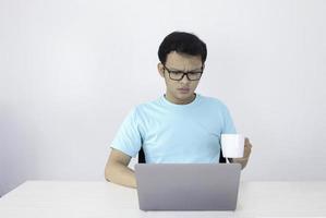 jovem asiático é sério quando trabalha no laptop com bebida café em vidro branco. homem indonésia usar fundo cinza isolado de camisa azul. foto