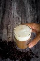 hora do café e conceito de refresco. mão segurando um copo transparente de café com espuma de leite e grão de café torrado em fundo de madeira.