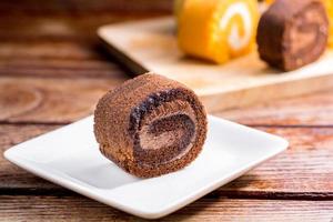 Feche o delicioso bolo de rolo de chocolate com chantilly em um prato quadrado branco em uma mesa de madeira na cozinha de casa com um fundo preto e espaço de cópia. conceito de padaria caseira. foto