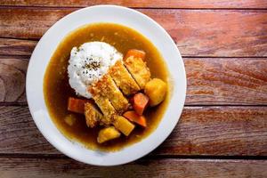 caril japonês com arroz, gergelim preto, cenoura, batata e frango frito em prato branco na mesa de madeira. foto