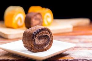 Feche o delicioso bolo de rolo de chocolate com chantilly em um prato quadrado branco em uma mesa de madeira na cozinha de casa com um fundo preto e espaço de cópia. conceito de padaria caseira. foto