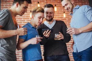 alegres velhos amigos se comunicam e observam o telefone, copos de uísque no pub. estilo de vida de entretenimento de conceito. pessoas conectadas por wifi em reunião de mesa de bar foto