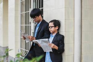 dois adolescente usando óculos, camisa azul e paletó preto encostado na parede e lendo jornal foto