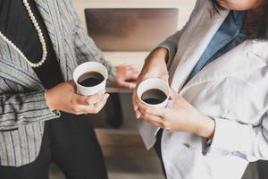 foto recortada de duas mulheres de negócios torradas em conquista de sucesso, duas trabalhadoras em pé e segurando uma xícara de café no escritório moderno, bem-sucedido, celebração, novo projeto, conceito de escritório de equipe