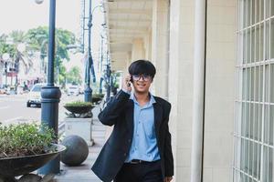 jovem asiático vestindo terno preto e óculos falar ao telefone enquanto sorrindo e andando foto