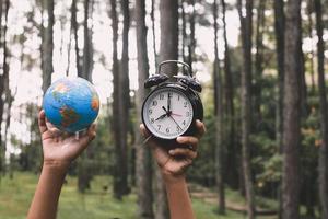 globo terrestre levantado à mão e despertador com embaçamento de árvores na floresta ao fundo, conceito de hora da terra foto