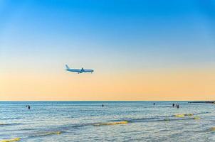 avião voando baixo acima do mar e turistas de pessoas nadando na água, céu azul claro e laranja ao pôr do sol, avião se preparando para pousar foto