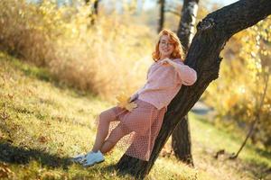 retratos de uma encantadora garota ruiva com um rosto bonito. garota posando no parque outono em um suéter e uma saia cor de coral. nas mãos de uma menina uma folha amarela foto