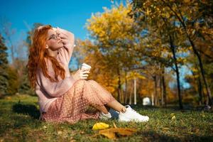 retratos de uma encantadora garota ruiva com óculos e um rosto bonito. garota posando no parque outono em um suéter e uma saia de cor coral. foto