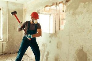 construtor com um martelo nas mãos quebra a parede de cimento o construtor está vestido com um traje de proteção e capacete foto