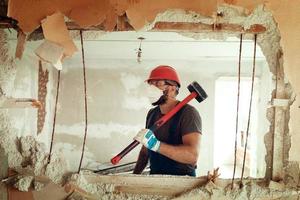 construtor com um martelo nas mãos quebra a parede de cimento o construtor está vestido com um traje de proteção e capacete foto