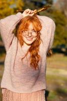 retratos de uma encantadora garota ruiva com óculos e rosto bonito. garota posando no parque outono em um suéter e uma saia cor de coral. a menina tem um humor maravilhoso foto