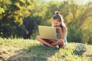 menina está falando em um laptop enquanto está sentado na grama ao sol. vestido com um sarafan e chapéu foto