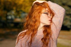 retratos de uma encantadora garota ruiva com óculos e rosto bonito. garota posando no parque outono em um suéter e uma saia cor de coral. a menina tem um humor maravilhoso foto