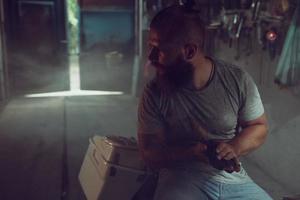 homem brutal bonito com barba sentado em uma motocicleta em sua garagem, limpando as mãos e olhando para o lado foto