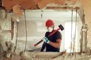 construtor com um martelo nas mãos quebra a parede de cimento o construtor está vestido com um traje de proteção e capacete foto