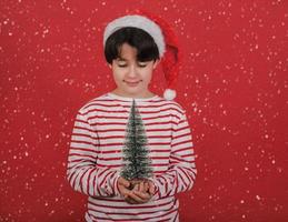 feliz natal, garoto usando chapéu de papai noel de natal com uma pequena árvore de natal nas mãos foto