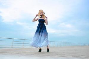 caucasiana linda loira elegante triste de vestido azul e chapéu de palha em pé à beira-mar foto