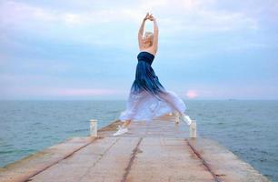 mulher loira branca caucasiana com cabelo curto em vestido longo e tênis feliz por estar grávida e pulando à beira-mar ao nascer do sol foto