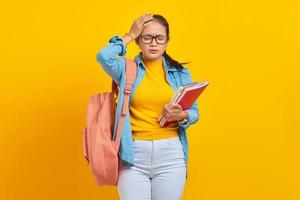 bela jovem estudante universitário asiático em roupa jeans com mochila segurando o notebook e parecendo tonto com dor de cabeça em fundo amarelo. educação no conceito de faculdade universitária do ensino médio foto