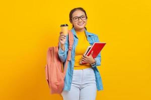 retrato de estudante asiática jovem alegre em roupas casuais com mochila segurando o livro e mostrando o café tirando olhando para câmera isolada em fundo amarelo