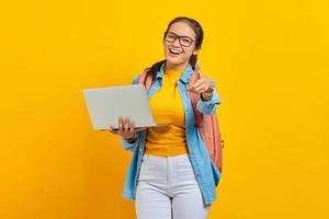 retrato de estudante asiática jovem alegre em roupas casuais com mochila usando laptop e apontando para a câmera com o dedo isolado no fundo amarelo. educação no conceito de universidade universitária foto