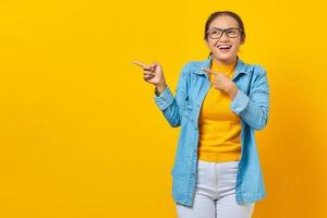 retrato de estudante asiática jovem alegre em roupas jeans, apontando para o espaço da cópia com o dedo e olhando de lado isolado no fundo amarelo. educação no conceito de universidade universitária foto