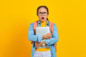 retrato de estudante surpreendida jovem mulher asiática em roupas casuais com mochila segurando laptop e olhando para câmera isolada em fundo amarelo. educação no conceito de faculdade universitária foto
