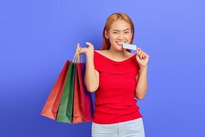 alegre linda mulher asiática de vestido vermelho segurando sacolas de compras e cartão de crédito isolado sobre fundo roxo foto