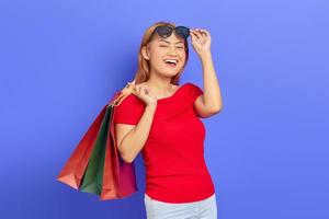 alegre linda mulher asiática de vestido vermelho e óculos segurando sacolas de compras isoladas sobre fundo roxo foto