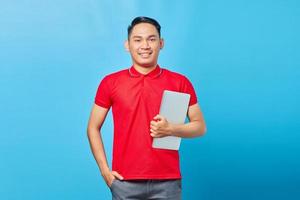 retrato de sorridente jovem bonito asiático de camisa vermelha segurando laptop e olhando para câmera isolada em fundo azul foto