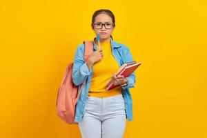 retrato de estudante séria jovem asiática em roupas casuais com mochila segurando livro e caneta, pensando em questão isolada em fundo amarelo. educação no conceito de universidade universitária foto