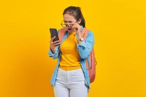 retrato de estudante asiática jovem surpresa em roupas casuais com mochila usando smartphone e tirando óculos isolados em fundo amarelo