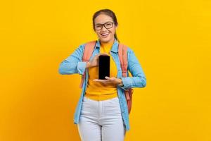 retrato de estudante asiática jovem alegre em roupas casuais com mochila mostrando celular de tela em branco isolado em fundo amarelo. educação no conceito de universidade universitária foto