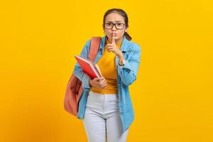 bela jovem estudante em roupas jeans com mochila e segurando o livro, dedos cobrindo os lábios, fazendo um gesto silencioso sobre fundo amarelo. educação no conceito de faculdade universitária do ensino médio foto